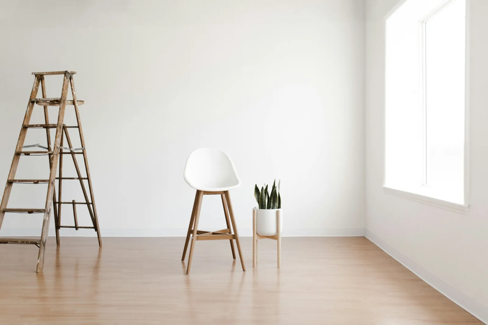 Pièce fraîchement rénovée aux murs blancs avec escabeau en bois, tabouret et plante près d’une fenêtre lumineuse